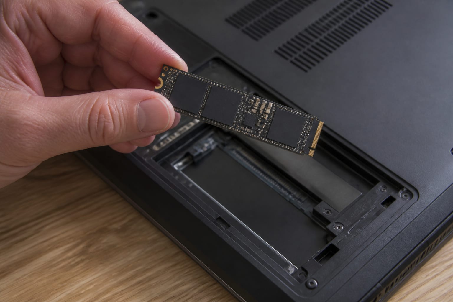 A hand holding an M.2 SSD above a laptop drive bay
