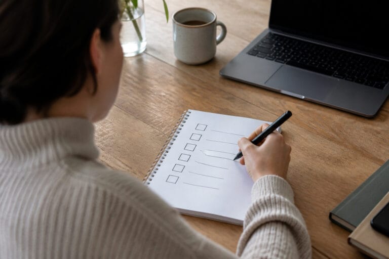 A person at a desk sketching a checklist next to a laptop